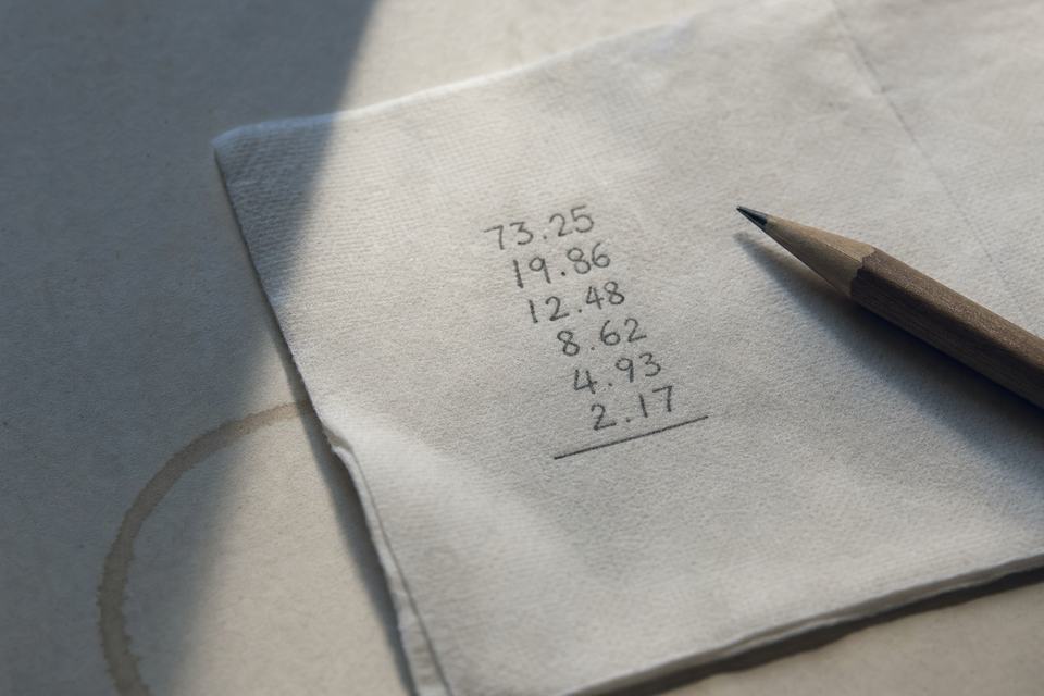 A paper napkin with six pencilled decimal figures in a column above a ruled line, a wooden pencil resting beside it and a faint cup ring on the desk.