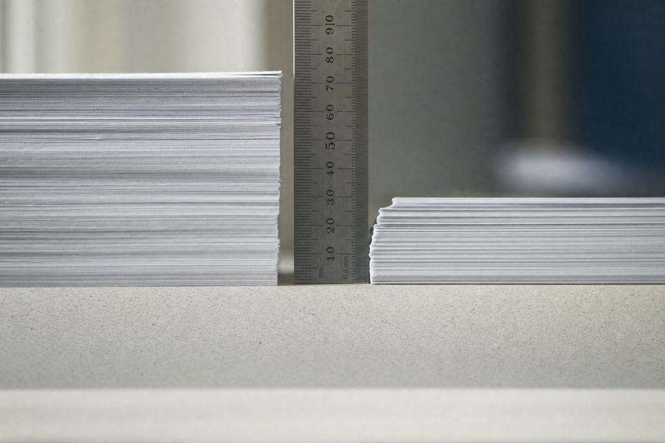 Two slabs of stacked paper of different heights in profile on a desk, a steel rule standing between them, morning light from the left.